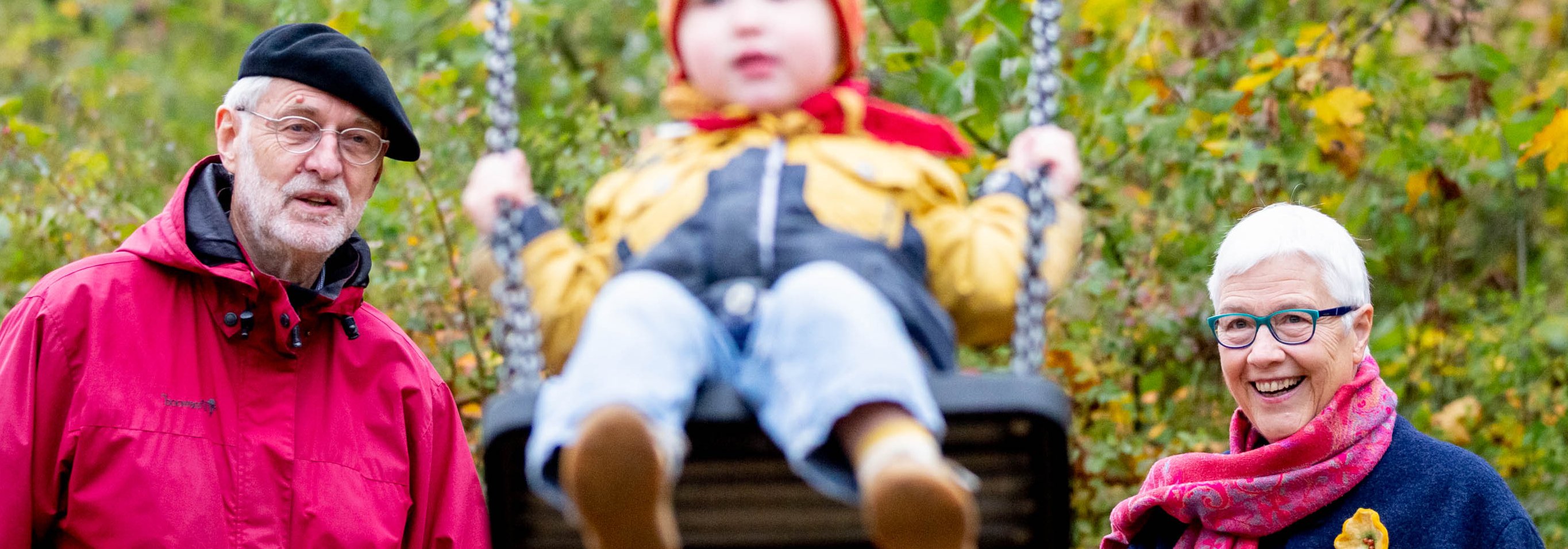 Das Foto zeigt ein &auml;lteres Paar, die hinter einer Schaukel stehen, auf der ein Kind sitzt.