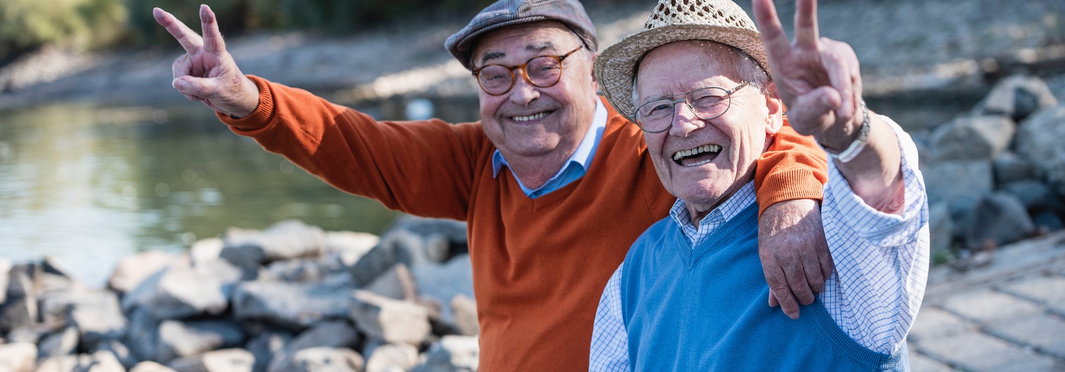 Zwei &auml;ltere M&auml;nner sitzen nebeneinander an einem liesigen Strand, sind gut gelaunt und machen Victory-Zeichen in Richtung des Fotografen