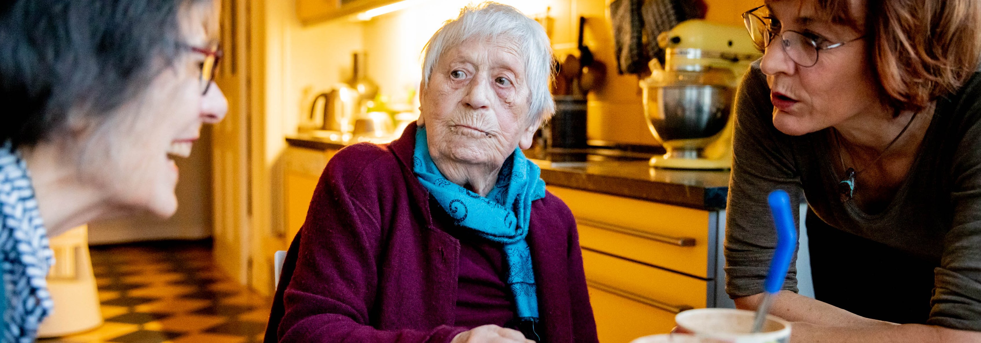 Das Foto zeigt drei Frauen an einem Tisch in einer K&uuml;che sitzend. Die mittlere Person ist &auml;lter als die beiden anderen, die sich scheinbar um sie k&uuml;mmern.